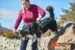 En kvinde hjælper sin hund, iført den grønne Non-stop Dogwear Ramble Harness, over en sten i naturen. Håndtaget på ryggen af selen gør det nemt at løfte og styre hunden i udfordrende terræn.